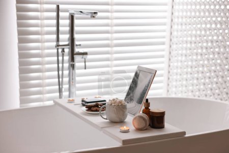 Téléchargez les photos : Plateau en bois blanc avec tablette, produits de spa, boisson chaude et bougie allumée sur la baignoire dans la salle de bain - en image libre de droit