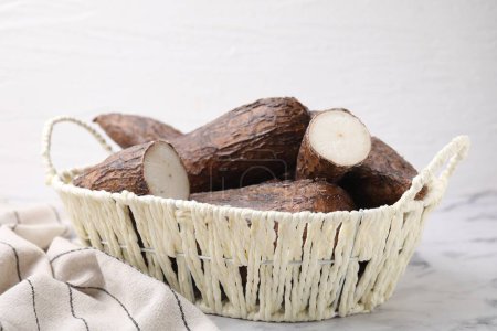 Téléchargez les photos : Racines de manioc entières et coupées dans un panier en osier sur une table en marbre blanc - en image libre de droit