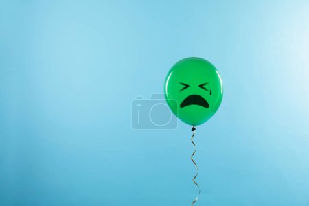 Green balloon with sad face on light blue background. Space for text