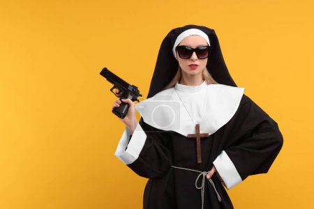 Photo for Woman in nun habit and sunglasses holding handgun against orange background, space for text - Royalty Free Image