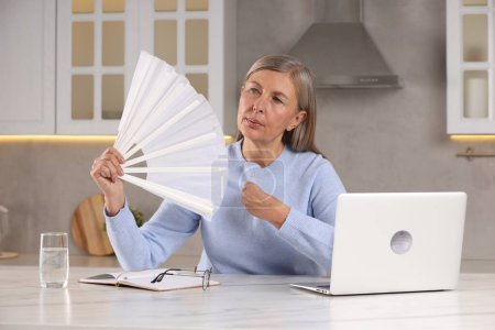 Photo for Menopause. Woman waving hand fan to cool herself during hot flash at table in kitchen - Royalty Free Image