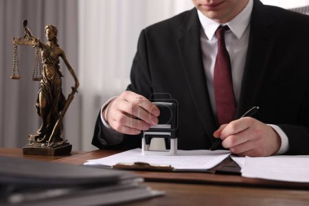 Notary with pen stamping document at wooden table in office, closeup