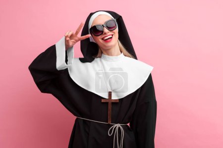 Photo for Happy woman in nun habit and sunglasses showing V-sign against pink background - Royalty Free Image