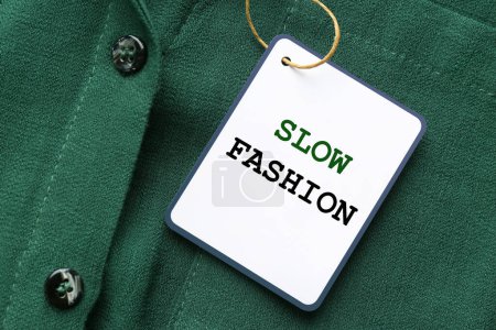 Conscious consumption. Tag with words Slow Fashion on green shirt, top view