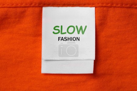 Bewusster Konsum. Bekleidungsetikett mit Schriftzug Slow Fashion auf orangefarbenem Kleidungsstück, Ansicht von oben