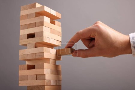 Playing Jenga. Man removing wooden block from tower on grey background, closeup