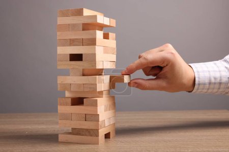 Playing Jenga. Man removing block from tower at wooden table against grey background, closeup