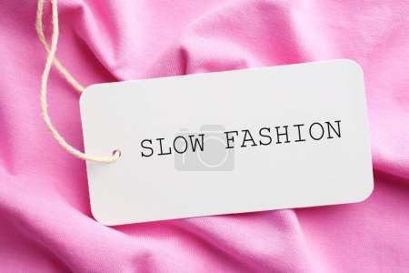 Conscious consumption. Tag with words Slow Fashion on pink fabric, top view