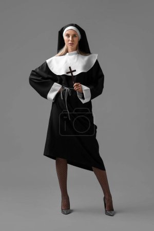 Woman in nun habit and mesh tights holding wooden cross on grey background. Sexy costume