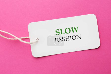 Conscious consumption. Tag with words Slow Fashion on pink background, top view