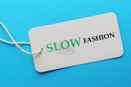 Conscious consumption. Tag with words Slow Fashion on light blue background, top view