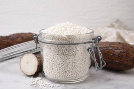 Téléchargez les photos : Perles de tapioca en pot et racines de manioc sur table blanche - en image libre de droit