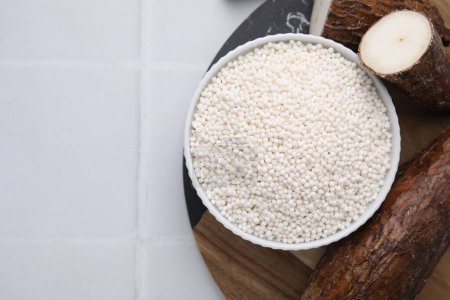 Téléchargez les photos : Perles de tapioca dans un bol et racines de manioc sur table carrelée blanche, plat. Espace pour le texte - en image libre de droit