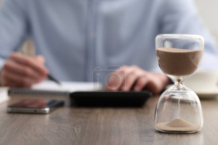 Hourglass with flowing sand on desk. Man taking notes while using calculator indoors, selective focus