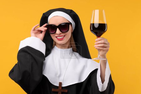 Happy woman in nun habit and sunglasses holding glass of wine against orange background. Sexy costume