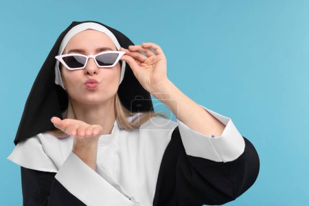 Woman in nun habit and sunglasses blowing kiss against light blue background. Space for text