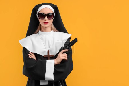 Femme en habit de religieuse et lunettes de soleil tenant une arme de poing sur fond orange, espace pour le texte