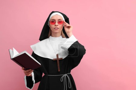 Mujer con hábito de monja y gafas de sol sosteniendo la Biblia sobre fondo rosa. Espacio para texto