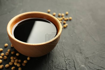 Tasty soy sauce in bowl and soybeans on black table, closeup. Space for text