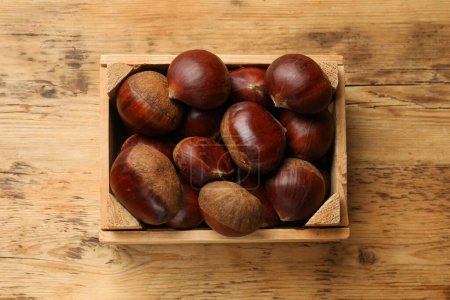Sweet fresh edible chestnuts in crate on wooden table, top view