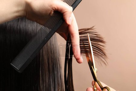 Hairdresser cutting client's hair with scissors on light brown background, closeup