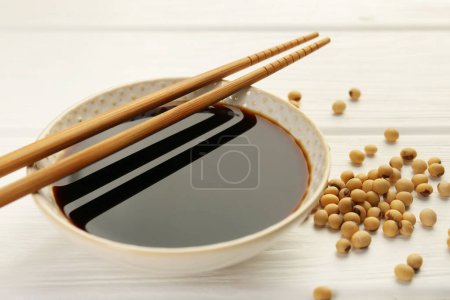 Tasty soy sauce in bowl, chopsticks and soybeans on white wooden table, closeup