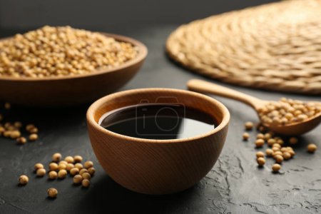 Tasty soy sauce in bowl and soybeans on black table, closeup