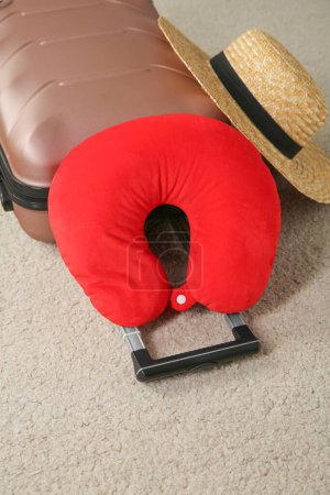 Red travel pillow, suitcase and hat on beige rug, above view