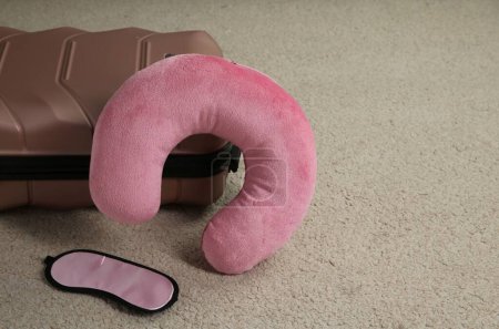 Pink travel pillow, suitcase and sleep mask on beige rug, space for text