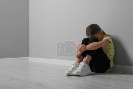 Abuso infantil. Muchacha molesta sentada en el suelo cerca de la pared gris, espacio para el texto