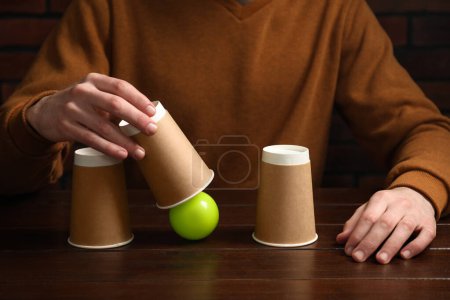 Muschelspiel. Mann zeigt Ball unter Becher an Holztisch, Nahaufnahme