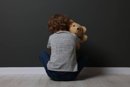 Abuso infantil. Niño trastornado con juguete sentado en el suelo cerca de la pared gris, vista trasera