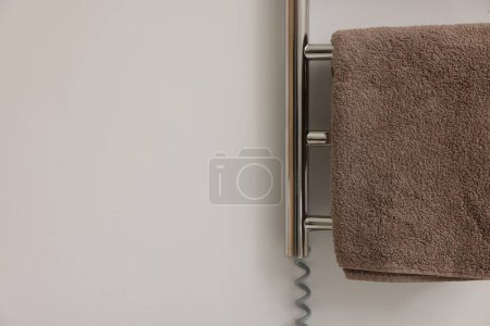 Heated rail with brown towel on white wall, closeup. Space for text