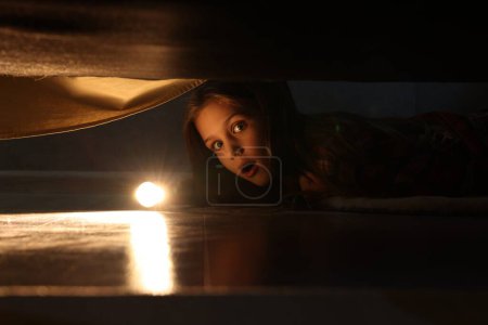 Téléchargez les photos : Petite fille avec lampe de poche à la recherche de monstre sous le lit la nuit - en image libre de droit