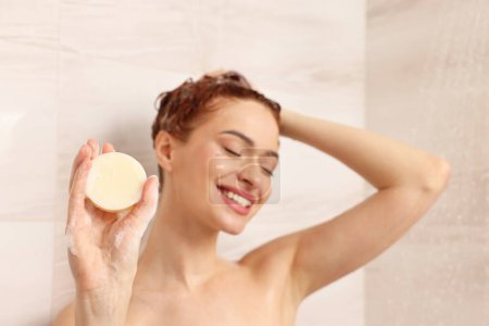 Happy young woman with solid shampoo bar in shower, selective focus