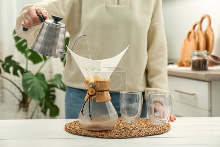 Frau gießt heißes Wasser in Glas Chemex-Kaffeemaschine mit Papierfilter und Kaffee an weißem Tisch, Nahaufnahme