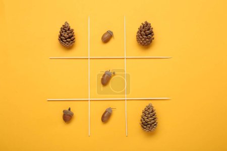 Tic tac toe game made with acorns and pine cones on yellow background, top view
