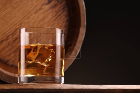 Whisky con cubitos de hielo en vidrio y barril sobre mesa de madera sobre fondo negro, espacio para texto