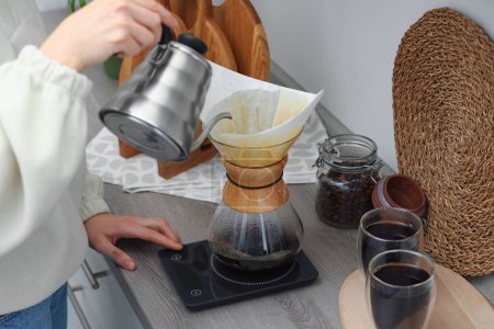 Frau gießt heißes Wasser in Glas Chemex-Kaffeemaschine mit Papierfilter und Kaffee an Arbeitsplatte in Küche, Nahaufnahme