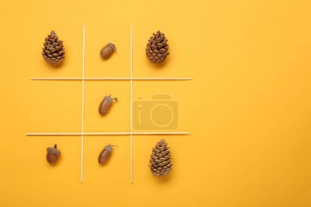 Tic tac toe game made with acorns and pine cones on yellow background, top view. Space for text