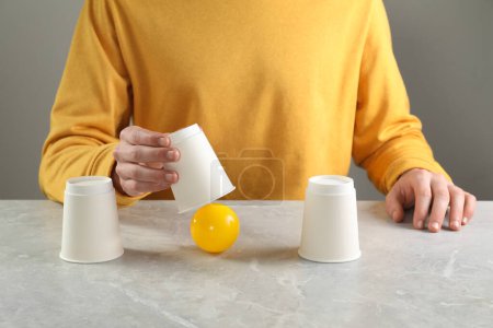 Shell game. Man showing ball under cup at light marble table, closeup