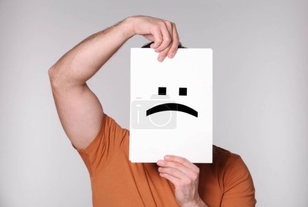 Man hiding behind sheet of paper with sad face on grey background