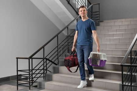 Travel with pet. Man holding carrier with cute cat and bag on stairs indoors