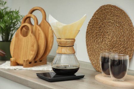 Photo for Glass chemex coffeemaker with paper filter, scales and glasses of coffee on wooden countertop in kitchen - Royalty Free Image