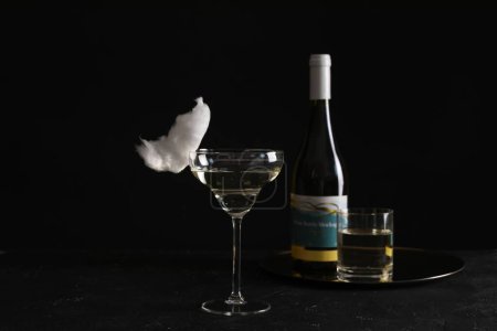 Cocktail with tasty cotton candy and bottle of alcohol drink on dark textured table against black background