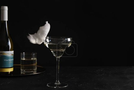 Cocktail with tasty cotton candy and bottle of alcohol drink on dark textured table against black background, space for text