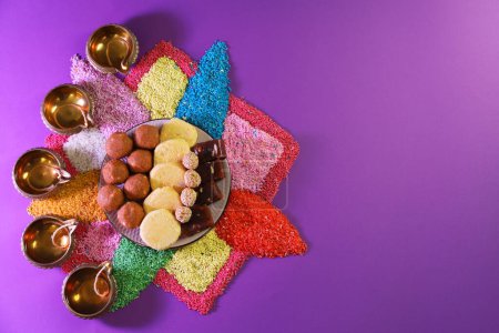 Diwali celebration. Tasty Indian sweets, colorful rangoli and diya lamps on violet background, flat lay. Space for text