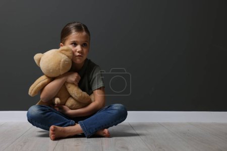 Kindesmissbrauch. Verärgertes Mädchen mit Spielzeug sitzt auf dem Boden neben grauer Wand, Platz für Text