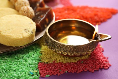 Diwali-Feier. Leckere indische Süßigkeiten, Diya-Lampe und bunte Rangoli auf violettem Tisch, Nahaufnahme