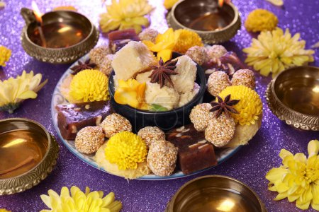 Diwali celebration. Diya lamps, tasty Indian sweets and yellow flowers on shiny violet table